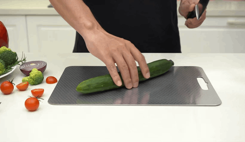 Hands holding a cucumber on a dark metal cutting board in a home kitchen.